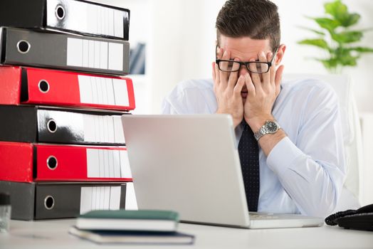 Young worried Businessman sitting in the office with hands on his face.  In front of his a bunch of binders.