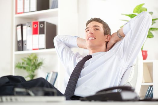Young successful businessman relaxing in the office with hands behind  head and daydreaming. In the background you can see the shelves with binders.