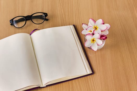 Plumeria or frangipani flowers in glass with blank note book and fresh relax mood, empty note book with frangipani or plumeria flowers on work table for copy space area, blank page diary on wooden table