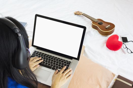Asian black hair girl sitting on bed surfing internet or working on notebook or laptop and listening to music with wireless headphone in relax mood with background of ukulele, eyeglasses,smartphone and red heart
