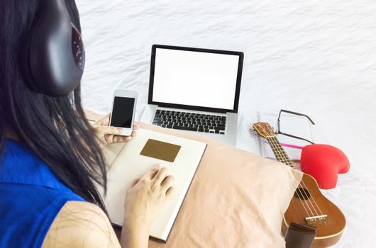 Blank laptop screen with Asian girl sitting on bed surfing internet and listening to music with wireless headphone, blank screen smartphone and empty card on note book,women buying music online