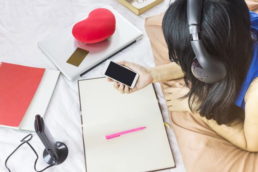 Black hair Asian girl lay down on bed surfing internet or shopping on website via smartphone with things around for background,women waiting phone call, women watching media on smartphone with blank note book and pen background