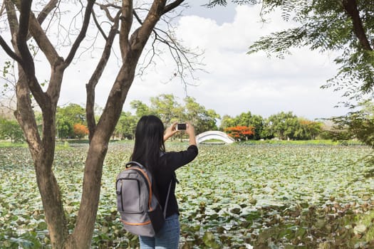 Back or rare view of girl or women shooting photo or video via smartphone beside the pond, girl or women in relax sunny puffy clouds blue sky day with smartphone in hands,girl with backpack and smartphone