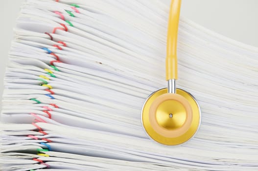 Yellow and gold stethoscope have blur pile paperwork of report with colorful paperclip as background.