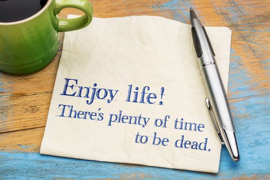 Enjoy life! There is plenty of time to be dead. Handwriting on a napkin with a cup of coffee,