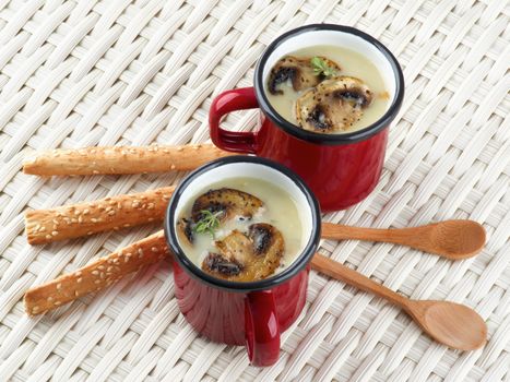 Arrangement of Delicious Homemade Mushrooms Cream Soup Decorated with Roasted Champignons in Red Cups with Wooden Spoons and Sesame Bread Sticks closeup on Wicker background