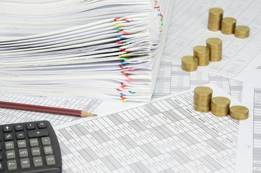 Brown pencil and step pile of gold coins on finance account have blur calculator and pile paperwork of report with colorful paperclip as background.