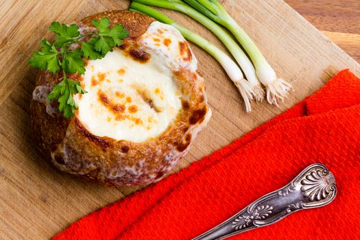 Tasty hot creamy French onion soup served in a bread bowl garnished with fresh parsley with a colorful red napkin and fresh spring onion ingredients on a wooden board