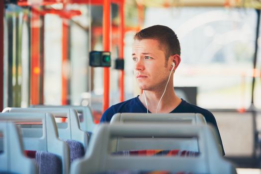 Sad young man is traveling by tram (bus). Everyday life and commuting to work by public transportation. Man is wearing headphones and listening to music.