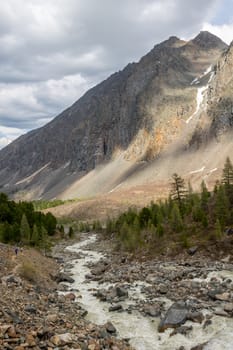 The mountain river, against the woody mountains, originating from a thawing glacier. Western Siberia, Altai mountains.
