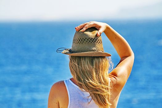 A young woman thoughtfully looks at a sea and holds a hat, hair develop on wind