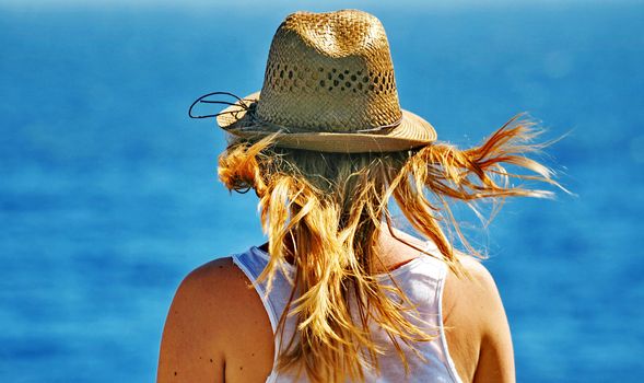 A young woman thoughtfully looks at a sea and holds a hat, hair develop on wind