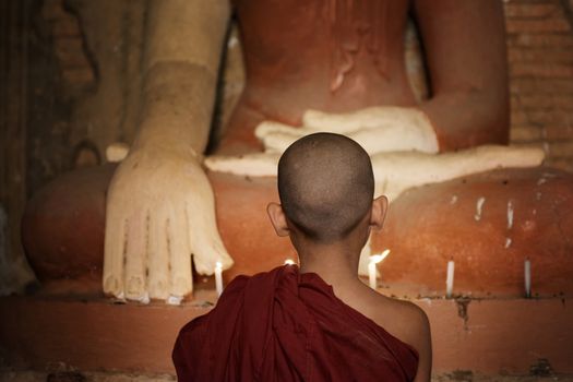 Rear view of young novice monk praying with candlelight inside a Buddhist temple, Bagan, Myanmar.