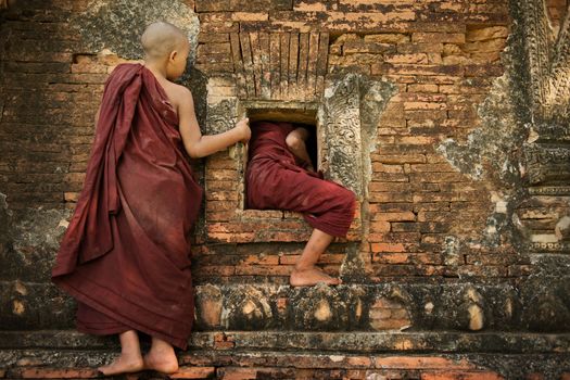 Two playful young novice monks climbing into Buddhist temple from window, Bagan, Myanmar.