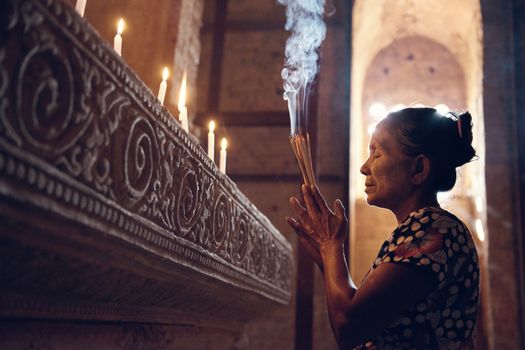 Old wrinkled traditional Asian woman praying with incense sticks inside a temple, low light, Myanmar