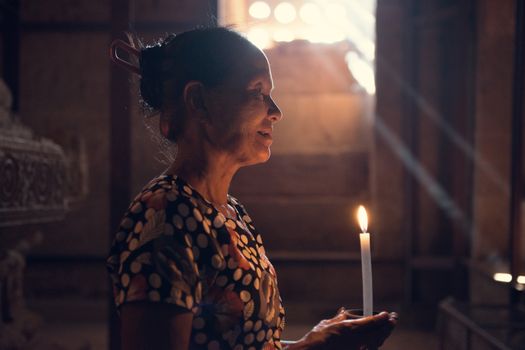 Old wrinkled traditional Asian burmese woman praying with candle light inside a temple, low light, Myanmar