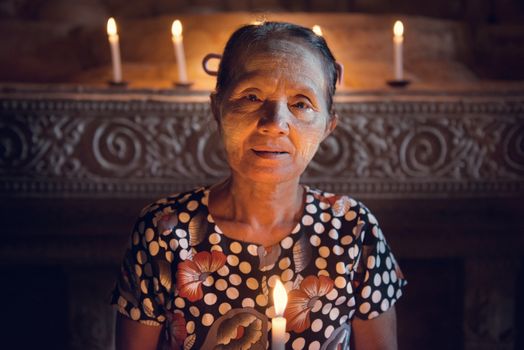 Old wrinkled traditional Asian woman praying with candle light inside a temple, low light, Myanmar