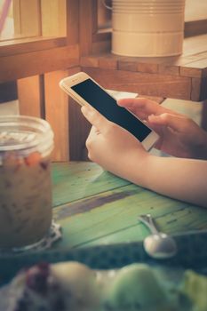 Woman hand holding smartphone on wood table in ice cream cafe on weekend afternoon. Trendy lifestyle concept with communication technology with vintage filter effect