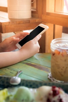 Woman hand holding smartphone on wood table in ice cream cafe on weekend afternoon. Trendy lifestyle concept with communication technology.