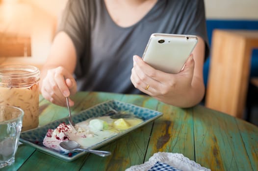 Young woman hand holding smartphone for read something while another hand get ice cream from dish in cafe on weekend afternoon. Modern lifestyle concept with technology