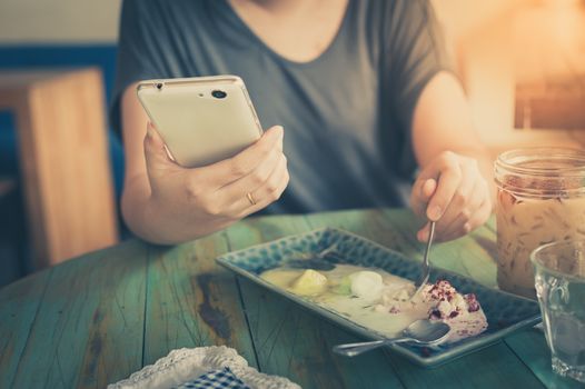 Young woman hand holding smartphone for read something while another hand get ice cream from dish in cafe on weekend afternoon. Modern lifestyle concept with technology. processed with vintage filter effect