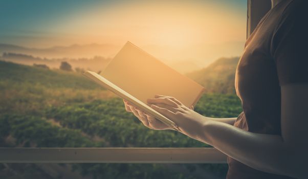 Young woman reading blank book beside window in morning time on weekend with mountain view in blurry background. Weekend lifestyle concept with vintage filter effect