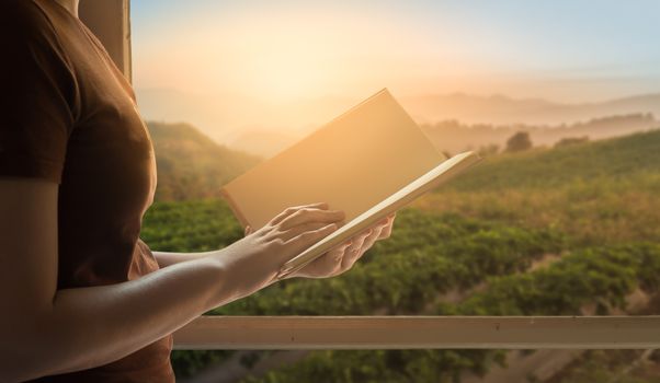 Young woman reading blank book beside window in morning time on weekend with mountain view in blurry background. Weekend lifestyle concept.