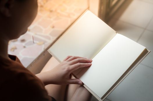 Young woman reading blank book while sitting beside window in morning time on weekend. Morning lifestyle on weekend concept.