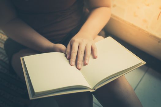 Young woman reading blank book while sitting beside window in morning time on weekend. Morning lifestyle on weekend concept with vintage filter effect