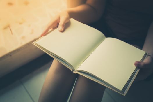 Young woman reading blank book while sitting beside window in morning time on weekend. Morning lifestyle on weekend concept with vintage filter effect