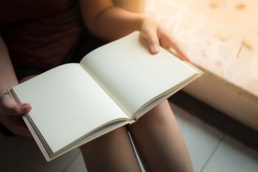Young woman reading blank book while sitting beside window in morning time on weekend. Morning lifestyle on weekend concept.