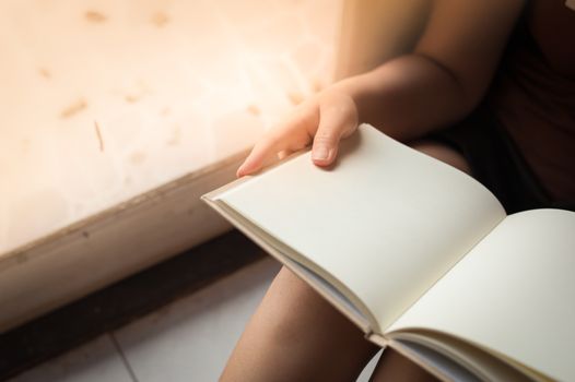 Young woman reading blank book while sitting beside window in morning time on weekend. Morning lifestyle on weekend concept.