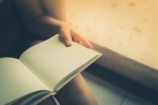 Young woman reading blank book while sitting beside window in morning time on weekend. Morning lifestyle on weekend concept with vintage filter effect