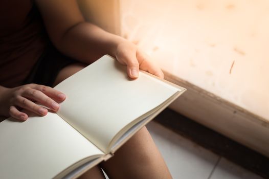 Young woman reading blank book while sitting beside window in morning time on weekend. Morning lifestyle on weekend concept.