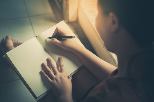 Young woman writing on blank book while sitting beside window in morning time on weekend. Morning lifestyle on weekend concept with vintage filter effect