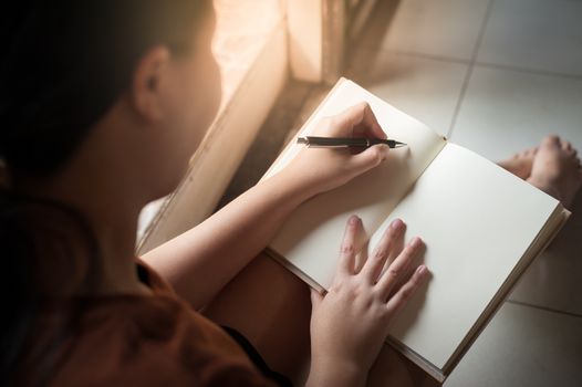 Young woman writing on blank book while sitting beside window in morning time on weekend. Morning lifestyle on weekend concept.