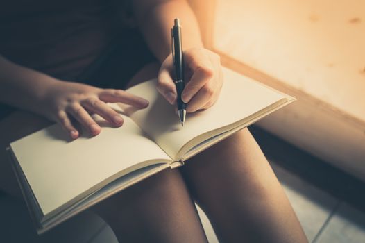 Young woman left hand writing on blank book while sitting beside window in morning time on weekend. Morning lifestyle on weekend concept with vintage filter effect