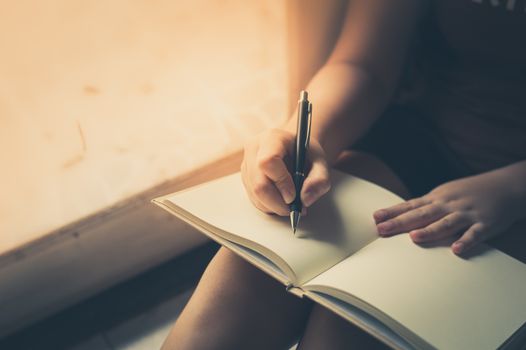 Young woman writing on blank book while sitting beside window in morning time on weekend. Morning lifestyle on weekend concept with vintage filter effect