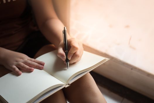 Young woman left hand writing on blank book while sitting beside window in morning time on weekend. Morning lifestyle on weekend concept.