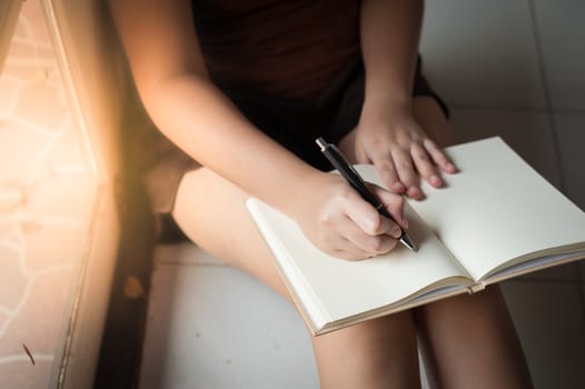 Young woman writing on blank book while sitting beside window in morning time on weekend. Morning lifestyle on weekend concept.