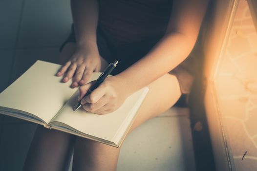 Young woman left hand writing on blank book while sitting beside window in morning time on weekend. Morning lifestyle on weekend concept with vintage filter effect