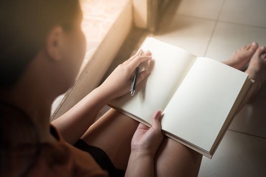 Young woman reading blank book while sitting beside window in morning time on weekend. Morning lifestyle on weekend concept.