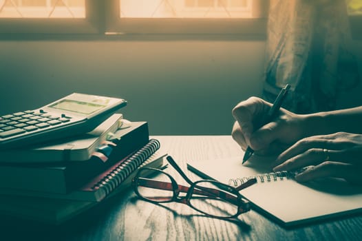 Male right hand writing on notebook beside glasses and books on table in morning time on work day. Freelance business working concpet with vintage filter effect