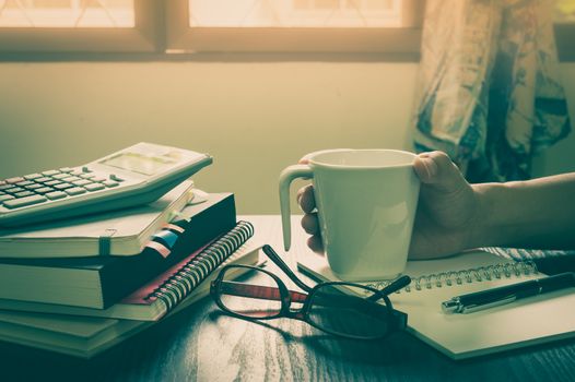 Male hand holding coffee cup on table beside notebooks, glasses, and pen in morning time on work day. Freelance business working concept with vintage filter effect