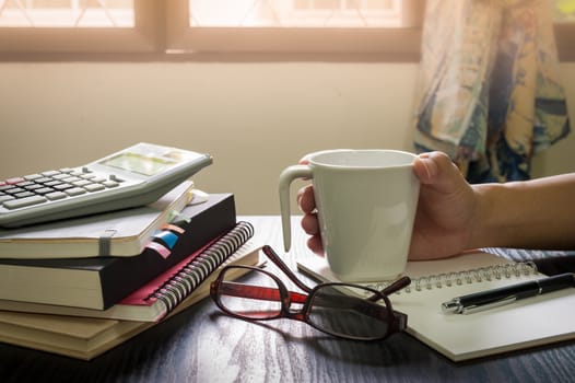 Male hand holding coffee cup on table beside notebooks, glasses, and pen in morning time on work day. Freelance business working concept.