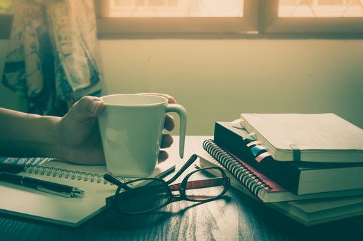 Young man left hand holding coffee cup on table beside notebooks, glasses, and pen in morning time on work day. Freelance business working concept with vintage filter effect