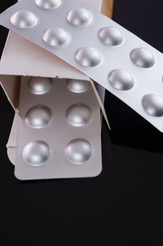 Silver blister pack of small pills together with the a box with additional packs showing through the open lid over a black background in a healthcare concept