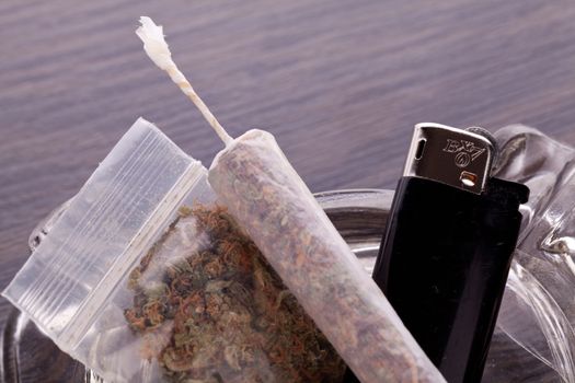 Close up of marijuana joint made with translucent rolling papers, plastic baggy of dried marijuana, black lighter and pipe on white background