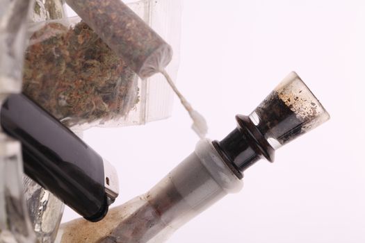 Close up of marijuana joint made with translucent rolling papers, plastic baggy of dried marijuana, black lighter and pipe on white background