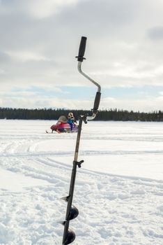 Scene with a ice fishing drill in the foreground while a parent and child is driving a snowmobile.